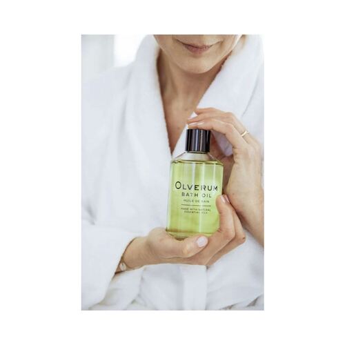Woman in white robe holding Olverum Bath Oil, emphasizing luxury self-care and aromatic body oil benefits.