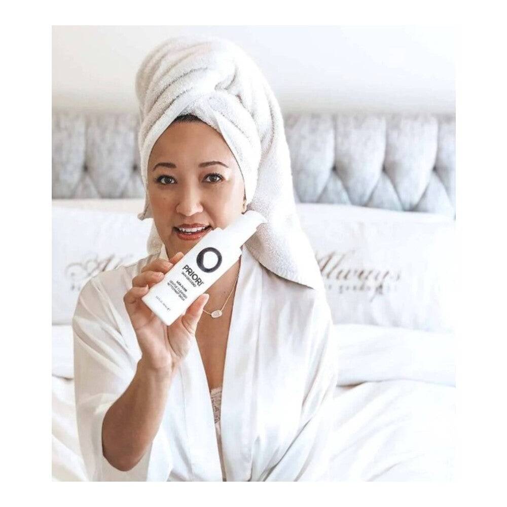 Woman in bathrobe holding Priori AHA Gentle Facial Cleanser, showcasing daily skincare use for radiant, smooth complexion.