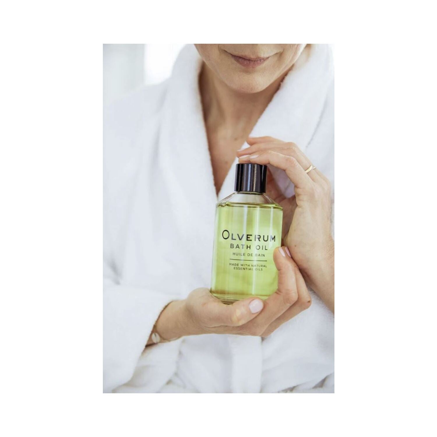 Person in white robe holding Olverum Bath Oil bottle. A calming body oil perfect for self-care and relaxation routines.