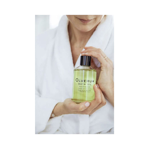 Person in white robe holding Olverum Bath Oil bottle. A calming body oil perfect for self-care and relaxation routines.