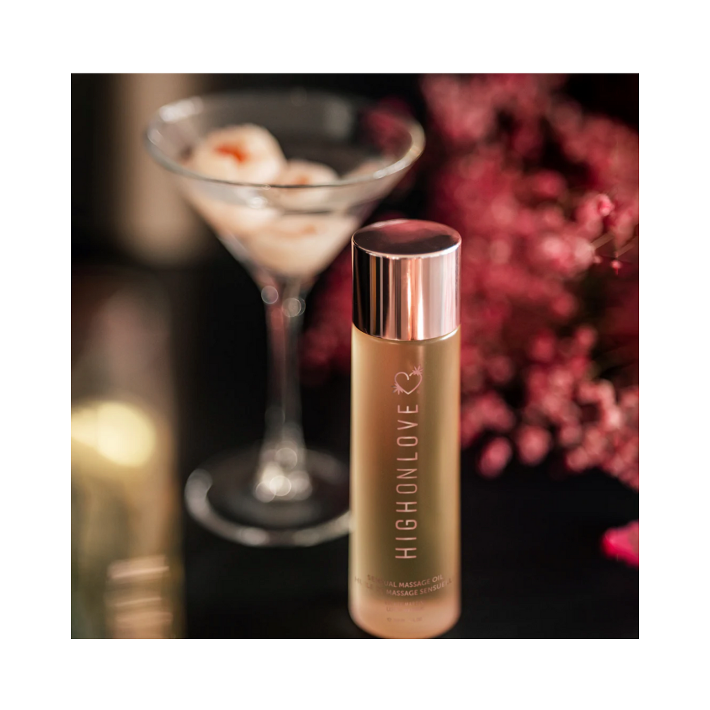 High on Love Massage Oil Topical Sunset Body Oil beside a martini glass and pink flowers, highlighting its romantic and luxurious appeal.