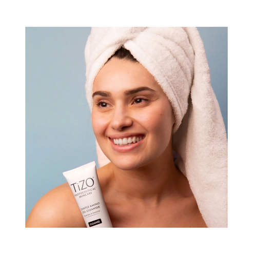 Smiling woman with towel holds TiZO Gentle Amino Cleanser. Promotes hydrated, radiant skin with soothing ingredients for daily facial care.